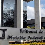 Fotografia em plano médio da fachada do Tribunal de Justiça do Distrito Federal e dos Territórios (TJDFT). Em primeiro plano, destaca-se um grande letreiro prateado com a sigla TJDFT em relevo escuro e a inscrição "Tribunal de Justiça do Distrito Federal e dos Territórios". Ao fundo, erguem-se pilares brancos e uma estrutura moderna com janelas espelhadas que refletem o céu com nuvens. Canteiros com arbustos verdes e flores amarelas ladeiam a base do monumento.