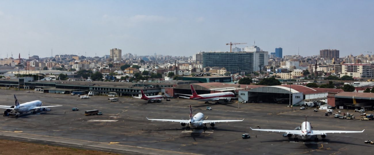 Vista panorâmica e aérea de uma placa de aeroporto com várias aeronaves comerciais de grande porte estacionadas na pista de manobra e junto aos hangares de manutenção. Em primeiro plano, destacam-se três aviões brancos alinhados. Ao fundo, estende-se uma densa paisagem urbana com edifícios residenciais e comerciais de média altura sob um céu claro com poucas nuvens, destacando-se uma grua de construção civil na linha do horizonte.