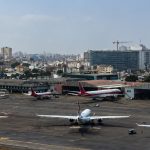 Vista panorâmica e aérea de uma placa de aeroporto com várias aeronaves comerciais de grande porte estacionadas na pista de manobra e junto aos hangares de manutenção. Em primeiro plano, destacam-se três aviões brancos alinhados. Ao fundo, estende-se uma densa paisagem urbana com edifícios residenciais e comerciais de média altura sob um céu claro com poucas nuvens, destacando-se uma grua de construção civil na linha do horizonte.