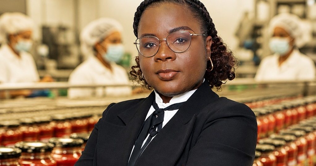 A imagem image_a7aba4.jpg mostra uma mulher africana de meia-idade em primeiro plano, com uma expressão séria e os braços cruzados. Ela tem cabelo escuro apanhado com tranças, usa óculos de armação fina e redonda, brincos de argola e veste um fato executivo preto com uma camisa branca e um laço escuro ao pescoço. Ela encontra-se no interior de uma unidade fabril, posicionada à frente de uma linha de produção repleta de frascos de vidro com polpa ou concentrado de tomate vermelho e tampas pretas. Ao fundo, três operárias com batas brancas, toucas e máscaras cirúrgicas cirulam desfocadas na zona de embalamento.