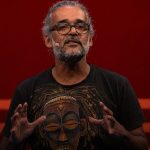 Fotografia em plano médio do activista angolano Luaty Beirão, de cabelos grisalhos apanhados, barba e óculos de massa escura, a discursar com as mãos erguidas no palco do Oslo Freedom Forum. Ele veste uma t-shirt preta com a ilustração detalhada de uma máscara africana colorida, contra um fundo vermelho uniforme.