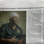 Fotografia focada na secção superior de uma página impressa do jornal norte-americano Amjambo Africa. O cabeçalho apresenta o título da peça "From hip-hop to resistance: Nelson Dembo’s fight for democracy in Angola". À esquerda, vê-se a imagem em plano médio do activista angolano Nelson Dembo, que usa uma touca de malha branca e uma camisa escura com um emblema circular no peito, surgindo em postura de diálogo com as mãos juntas. O texto jornalístico em inglês distribui-se em colunas ao lado e abaixo da fotografia.