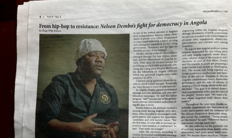Fotografia focada na secção superior de uma página impressa do jornal norte-americano Amjambo Africa. O cabeçalho apresenta o título da peça "From hip-hop to resistance: Nelson Dembo’s fight for democracy in Angola". À esquerda, vê-se a imagem em plano médio do activista angolano Nelson Dembo, que usa uma touca de malha branca e uma camisa escura com um emblema circular no peito, surgindo em postura de diálogo com as mãos juntas. O texto jornalístico em inglês distribui-se em colunas ao lado e abaixo da fotografia.