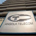 Fotografia em perspetiva de contra-plongé focando uma grande placa metálica retangular na entrada do edifício da Angola Telecom. A placa exibe o logótipo oficial da empresa, composto por um círculo estilizado com uma letra "T" estilizada no centro, acompanhado pela inscrição "ANGOLA TELECOM" em letras maiúsculas escuras na parte inferior. Ao fundo, ergue-se a fachada de um edifício residencial ou de escritórios com múltiplas varandas e janelas sob um céu claro.